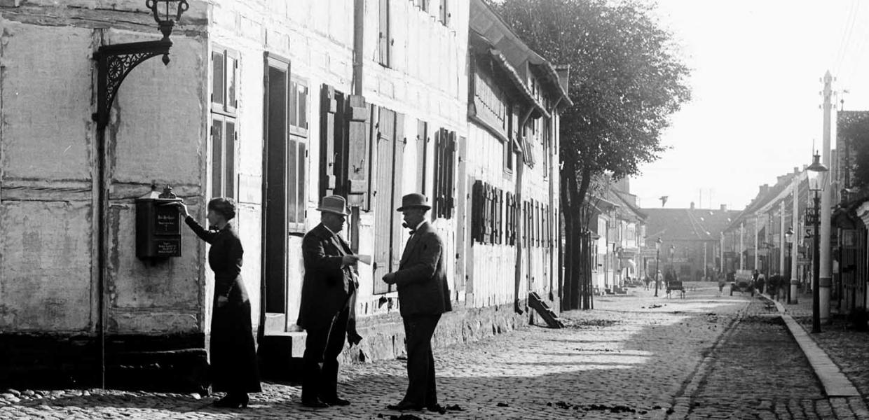A woman is standing to the side, and two men wearing bowler hats are talking together at Trægården in the olden days