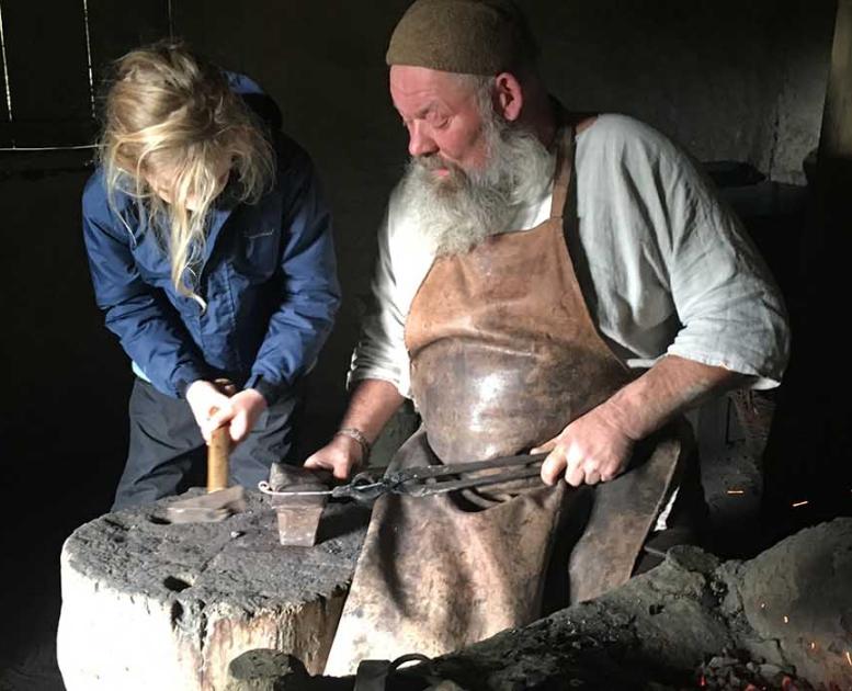 The Iron Age blacksmith holds the metal that the girl is to hammer on